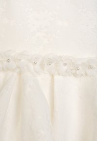 Weißes Kleid aus strukturiertem Blumenstoff, mit einem fliedernden Saum, der von einem Band aus Stoffblumen und kleinen Perlendetails akzentuiert wird.
