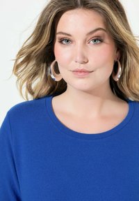 Maglione blu con scollo rotondo, morbido al tatto, abbinato a grandi orecchini a cerchio in argento. I capelli sono ondulati e cadono delicatamente intorno al viso.