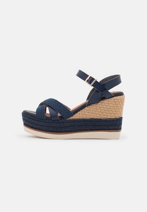 Marineblå kileformet sandal med flettede stropper, ankelspænde og vævet tekstureret hæl, der har en beige platformsål.
