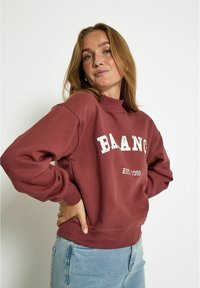 Sudadera burdeos con cuello redondo, presenta texto blanco "BAANC" y "EST.1999" en una fuente desgastada. Mangas largas, ajuste relajado y dobladillo acanalado.