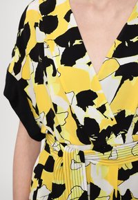 Femme portant une robe portefeuille à manches courtes avec un motif floral abstrait jaune, noir et blanc, nouée à la taille avec une ceinture assortie.