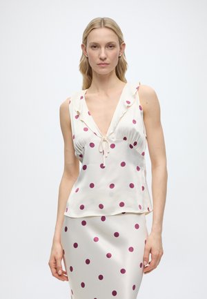 Kobieta w białym satynowym topie bez rękawów i spódnicy w fioletowe kropki, z głębokim dekoltem w serek i wiązaniem na topie.