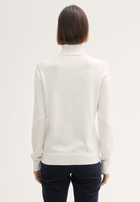 Žena s krátkými hnědými vlasy, oblečená v bílém roláku a tmavých kalhotách, zobrazená zezadu na jednobarevném pozadí.