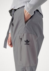 Grå träningsbyxor med en sidoficka med dragkedja och en broderad svart Adidas-logotyp. Tillverkade av lätt och smidigt material, med midja som kan justeras med dragsko.