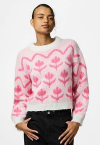 Maglione a maglia rosa e bianco con motivo floreale, scollo tondo e polsini a costine. La texture è morbida e soffice. Indossati jeans neri sotto.