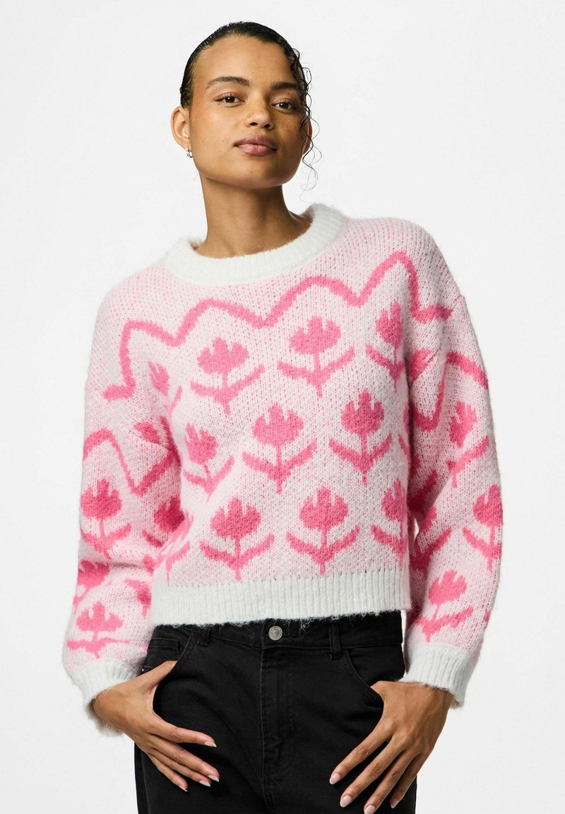 Maglione a maglia rosa e bianco con motivo floreale, scollo tondo e polsini a costine. La texture è morbida e soffice. Indossati jeans neri sotto.