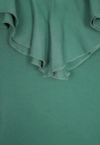 Grön textil med en volangkrage. Tyget är glatt med en svag lyster, vilket kompletteras av fina stygn längs kanterna.