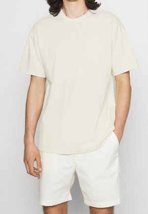 Mand iført cremefarvet kortærmet T-shirt og matchende shorts, stående med den ene hånd i lommen mod en ensfarvet baggrund.