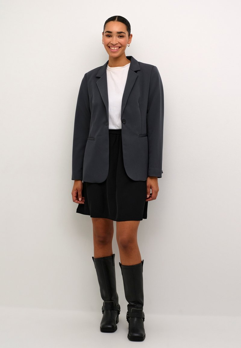 Blazer gris à un bouton, jupe noire et bottes en cuir noir jusqu'aux genoux. T-shirt blanc uni en dessous.