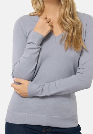 Kvinde iført en lysegrå V-hals sweater med lange ærmer og mørkeblå jeans, der holder den ene arm over taljen og rører ved halsudskæringen.