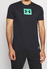 Svart kortärmad T-shirt i slät tyg, med en grön fyrkantig logotyp i mitten och en grön etikett på den nedre sidan.