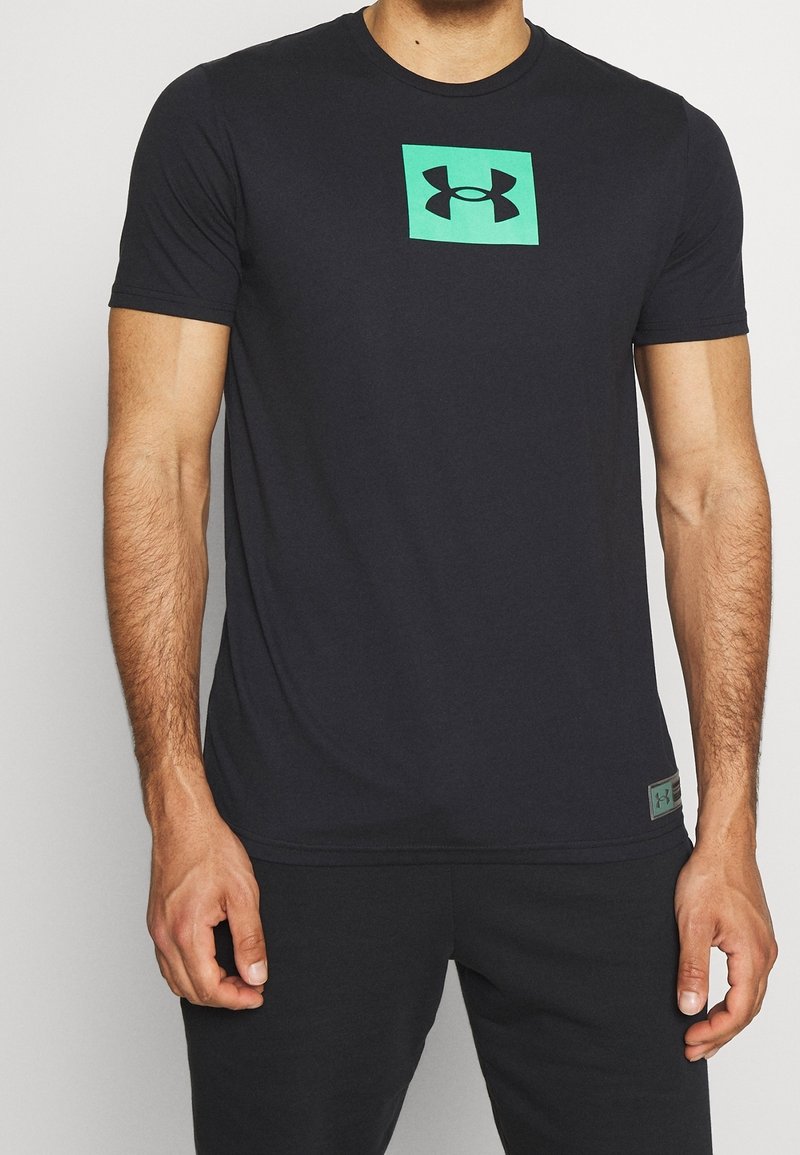 Svart kortärmad T-shirt i slät tyg, med en grön fyrkantig logotyp i mitten och en grön etikett på den nedre sidan.