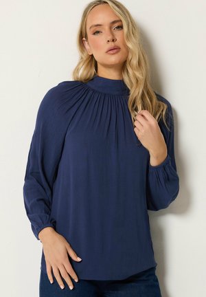 Frau mit langen blonden Haaren, die eine locker sitzende marineblaue Bluse mit langen Ärmeln und gerafftem Stoff am Ausschnitt und an den Manschetten trägt.