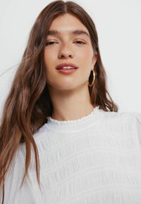 Bílá texturovaná blůza s volánkovým límcem, se svislými pruhy a měkkou tkaninou. Modelka má na sobě přirozený make-up a náušnice ve tvaru kruhu.