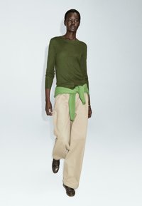 Maglione verde oliva a maniche lunghe con un maglione verde chiaro legato in vita, abbinato a pantaloni larghi beige e scarpe marroni.
