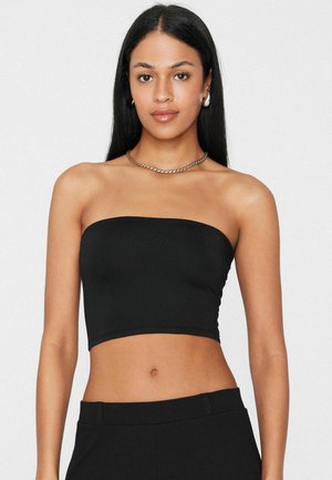 Junge Frau mit langen dunklen Haaren, die ein schwarzes trägerloses Crop-Top, schwarze Hose, silberne Halskette und silberne Creolen trägt.