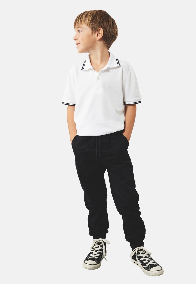 Polo bianca con colletto a righe bianche e nere, abbinata a pantaloni jogger neri. Sneakers nere con lacci e suole bianche.