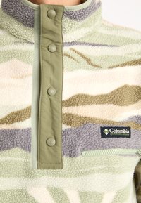 Pullover in pile con motivo camouflage verde, beige e viola. Presenta un colletto alto, bottoni a pressione e una tasca con zip dotata di un badge con logo.