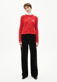 Roter Zopfmuster-Cardigan mit rundem Ausschnitt, zwei Fronttaschen und grauen Knöpfen. Kombiniert mit schwarzen weit geschnittenen Hosen und schwarzen Schuhen.