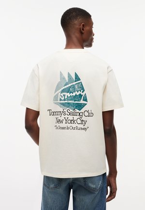 Bărbat purtând un tricou de culoare off-white cu grafică albastră a unei bărci cu vele și textul „Tommy's Sailing Club New York City” și „The Ocean Is Our Runway” pe spate.