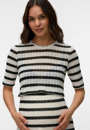 Kobieta z długimi ciemnymi włosami, ubrana w czarno-biały poziomo paskowany sweter oraz dopasowaną spódnicę na jednolitym tle.
