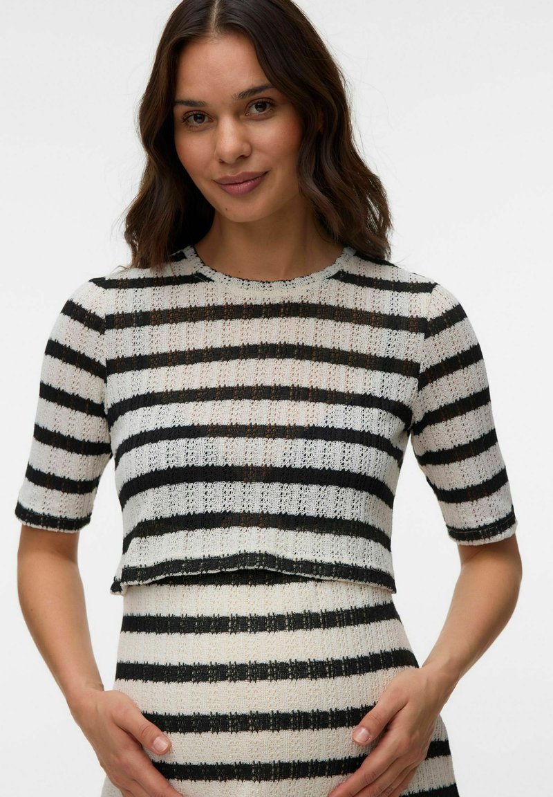 Femme aux longs cheveux foncés portant un haut tricoté à rayures horizontales noires et blanches et une jupe assortie sur un fond uni.
