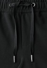 Nära bild av svart elastisk midjeband med svarta snören och silverfärgade metallspetsar på mörka tygbyxor.