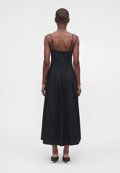 Personne aux cheveux courts portant une robe midi noire sans manches avec de fines bretelles et des sandales noires à talons, debout devant un fond uni.