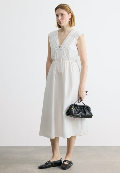 Femme portant une robe midi blanche sans manches avec un détail à cordon, tenant un sac à main noir plissé, et portant des ballerines noires pointues avec des détails de sangle.