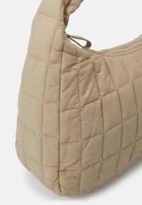 Quiltet beige skuldertaske med en afrundet form, der har synlige syninger og en tekstureret overflade. Indeholder en lynlåslukning øverst.