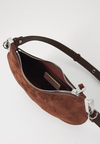 Borsa a tracolla in suede marrone con hardware argentato, chiusura a zip e una tracolla in pelle marrone. All'interno presenta una tasca piatta con un'etichetta.