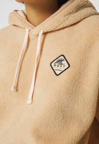 Cream fleece hoodie with a beige lining, featuring a black and yellow Roxy logo patch, drawstrings, and a soft texture.