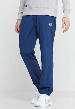 Mann trägt blaue Trainingshose mit elastischen Bündchen, ein hellgraues Sweatshirt und weiße Sportschuhe, steht vor einem einfarbigen Hintergrund.