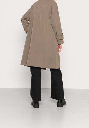 Person iført en løs taupe trenchcoat, sorte vidde bukser og sorte platåsko, set bagfra mod en ensartet lys baggrund.
