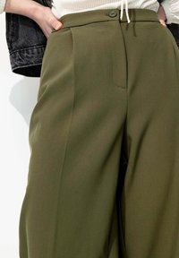 Pantalons ajustés vert olive à taille haute, avec un pli avant, une poche latérale et une fermeture à un bouton. Texture de tissu lisse.