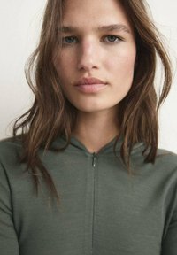 Zelena zip-up dukserica s mekanom teksturom, koja ima visoki ovratnik i jednostavan dizajn. Tkanina izgleda lagano i ležerno.