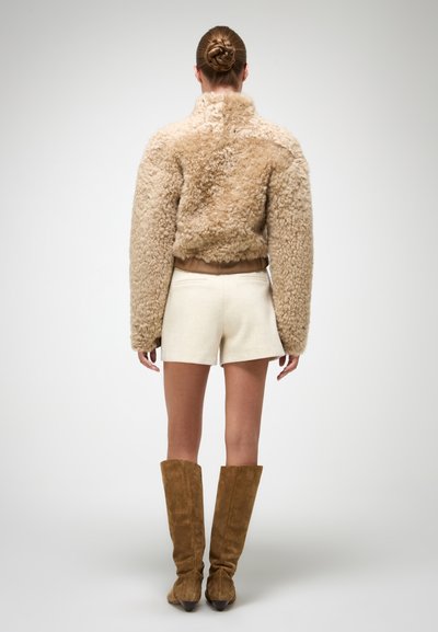 Béžová umelá kožušina s vysokým golierom, skrátený strih, nosená cez biele šortky a kombinovaná s vysokými hnedými semišovými čižmami. Hladká textura.