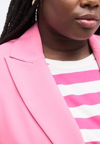 Γυναίκα με σκούρα πλεξούδες, φοράει ροζ σακάκι πάνω από λευκό και ροζ ριγέ πουκάμισο, τα χείλη κλειστά σε ουδέτερη έκφραση.