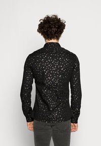 Camisa negra con un patrón de puntos dorados brillantes, manga larga y cuello. La tela tiene un acabado texturizado y brillante.