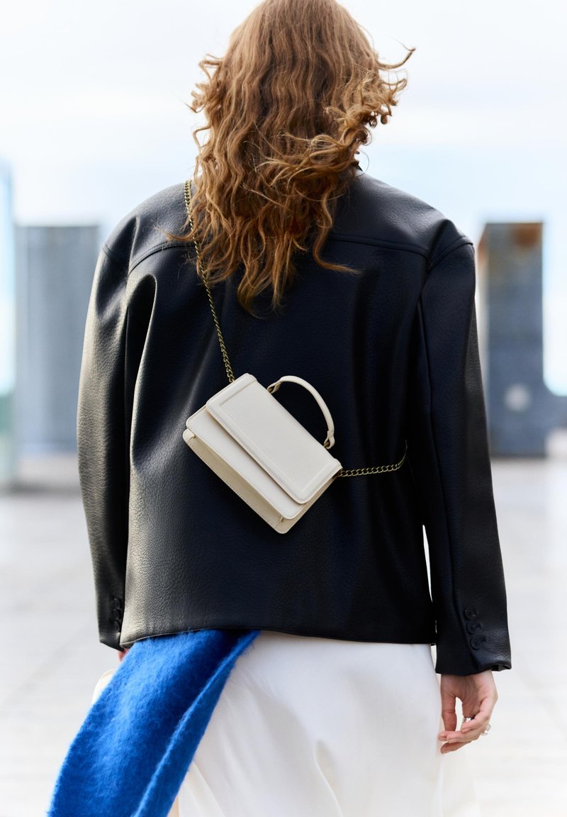 Personne aux longs cheveux bouclés portant une veste noire et une jupe blanche, portant un petit sac blanc à chaîne sur le dos en extérieur.