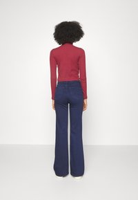 Paita turtleneck maroon yang dipadukan dengan celana jeans biru tua bermodel high-waisted dan flare yang dilengkapi saku belakang serta permukaan yang halus dan terstruktur.