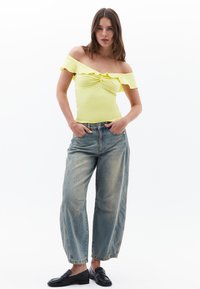 Top giallo scollato con bordo a volants; dettaglio frontale attorcigliato. Jeans a gamba larga azzurri con texture sbiadita; mocassini neri.