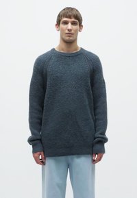 Dunkelgrauer strukturierter Pullover mit einem gerippten Rundhalsausschnitt und langen Ärmeln, versehen mit kontrastierenden Seitenpaneelen. Kombiniert mit hellblauen Hosen.
