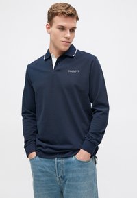 Jūras spēku garās piedurknes polo krekls ar baltām apkakles akcijām un "HACKETT" logo. Audums šķiet mīksts; brīva piegriezuma ar sānu šķelēm un ribotām piedurknēm.