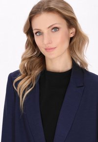 Námořnický modrý blazer s klopami nožících, přetažený přes černý top, vyrobený z texturované látky a s propracovaným střihem, předváděný osobou s vlnitými vlasy.