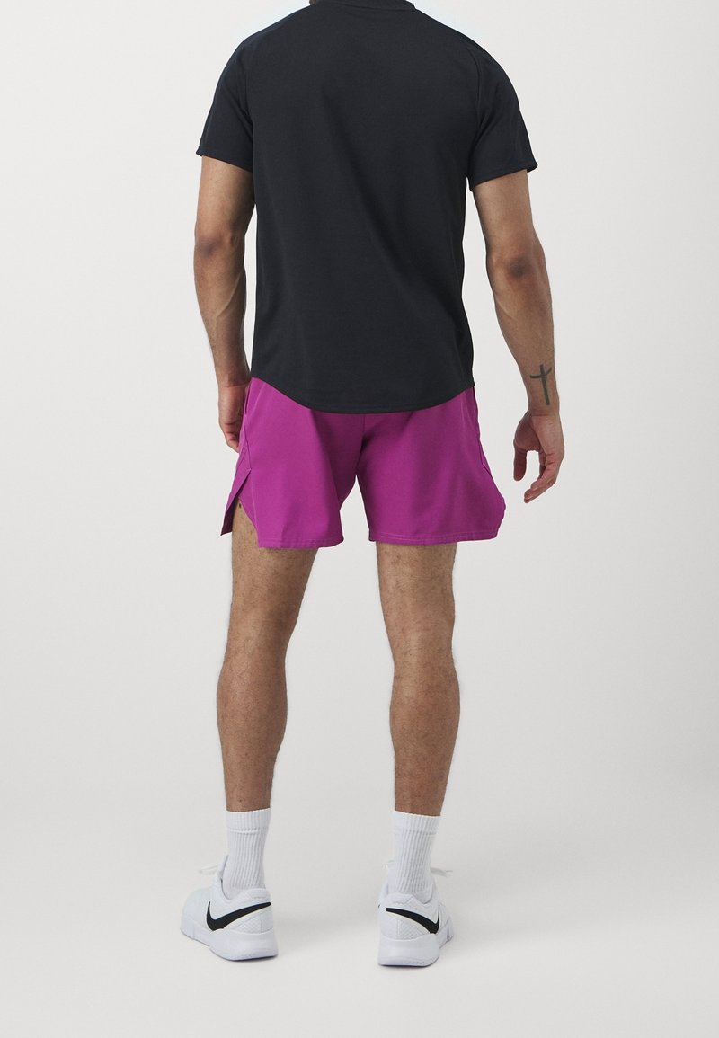 Homme portant une chemise noire à manches courtes, un short violet, des chaussettes blanches et des baskets blanches Nike, debout de dos sur un fond uni.