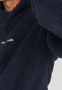 Granatowa bluza z polaru z fakturowaną, miękką powierzchnią i oversize'owymi rękawami. Biały napis "origin studio" na piersi.