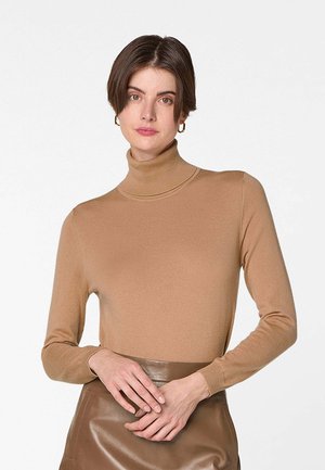 Vrouw met kort bruin haar, gouden creooloorbellen, een coltrui in kamelkleur en een bruine leren hoge taille rok.