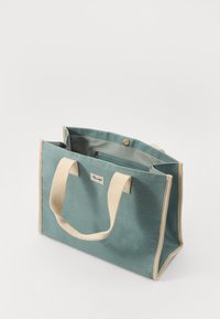 Lerret tote bag i teal med beige bomullshåndtak. Har en knappelukking, indre lomme, og et merke på framsiden.