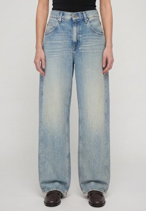 Femme aux longs cheveux blonds porte un haut noir sans manches, un jean large bleu clair et des chaussures noires, debout devant un fond uni.